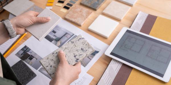 Hands of young female designer holding two samples of marble tile over wooden table with digital tablet, photos of home interior etc