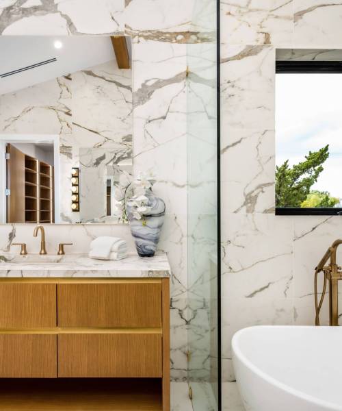 Modern bathroom interior with marble walls, wooden vanity, and gold fixtures, featuring a large mirror and a window with a view of greenery.